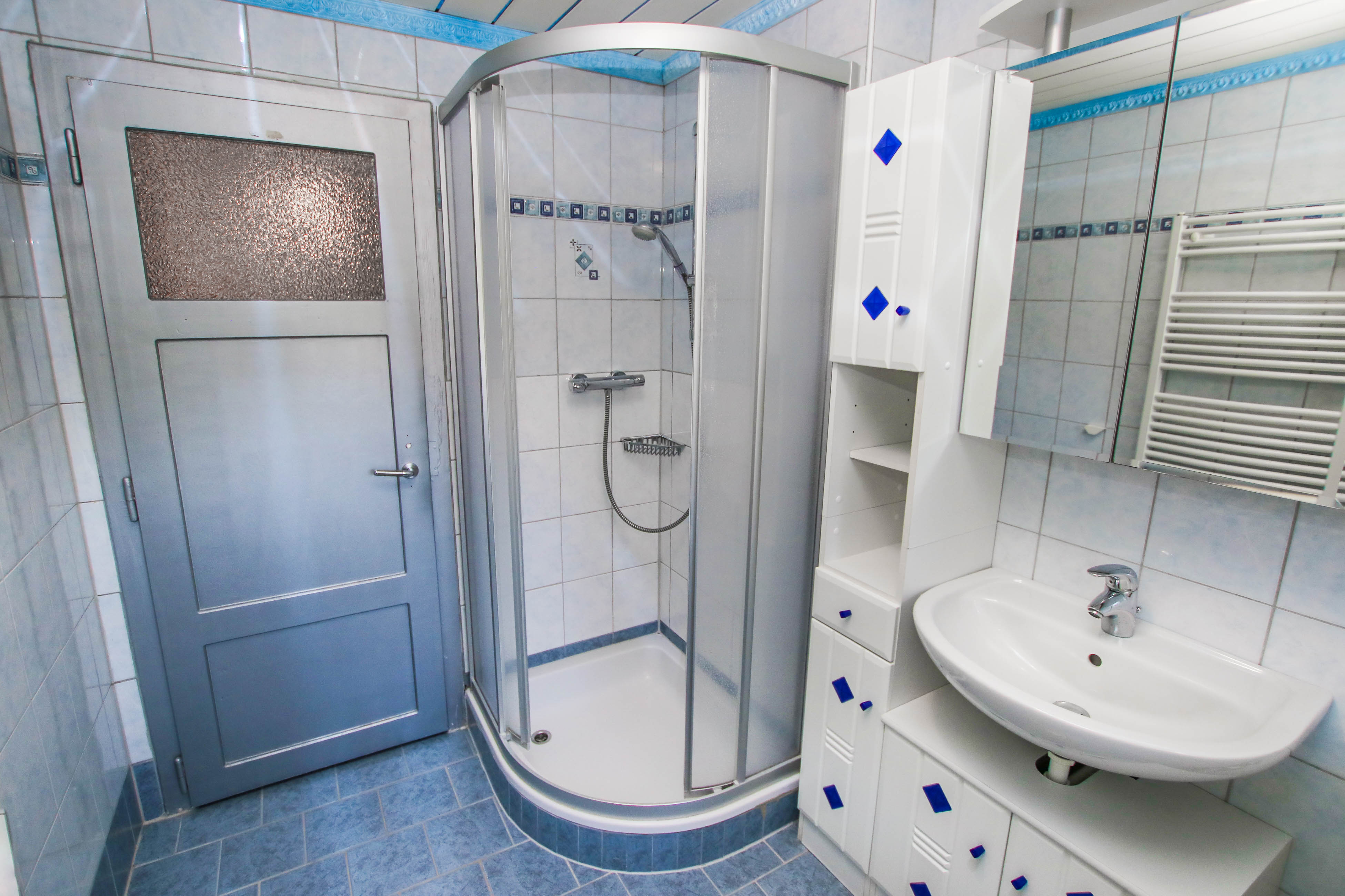 Das in weiß und blau gehaltene Badezimmer mit Blick auf die Dusche sowie das Waschbecken und das Badkästchen.